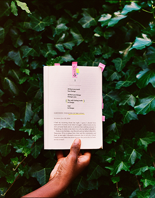 A hand holds an open, annotated book with colorful sticky tabs against a background of green ivy leaves.