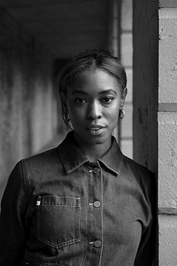 Black-and-white portrait of Jazmine. standing beside a concrete column, looking toward the camera in a buttoned denim jacket.