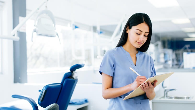 Female dental assistant in blue scrubs writing notes in a bright dental clinic with a dental chair in the background.